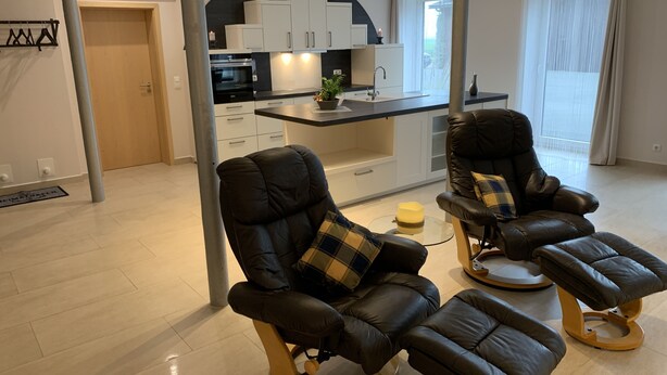 A modern living room with two leather recliners, a kitchen area with white cabinets, and a large window with curtains. - Apartment Ahsbahs Suite near Airport (Elskop)