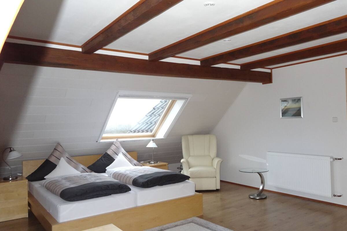 A bedroom with a sloped ceiling, wooden beams, a skylight, a bed with pillows, a chair, a small table, and a radiator.