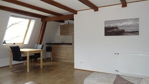 A room with a wooden dining table, chairs, a kitchen area with wooden cabinets, a radiator, and a painting of a landscape. - Apartment Panorama 2 near Airport (Elskop)