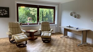 A room with two leather chairs, a small wooden table, a white desk, and a large window with a view of greenery. - Apartment Elbe Glückstadt near Airport (Glueckstadt)