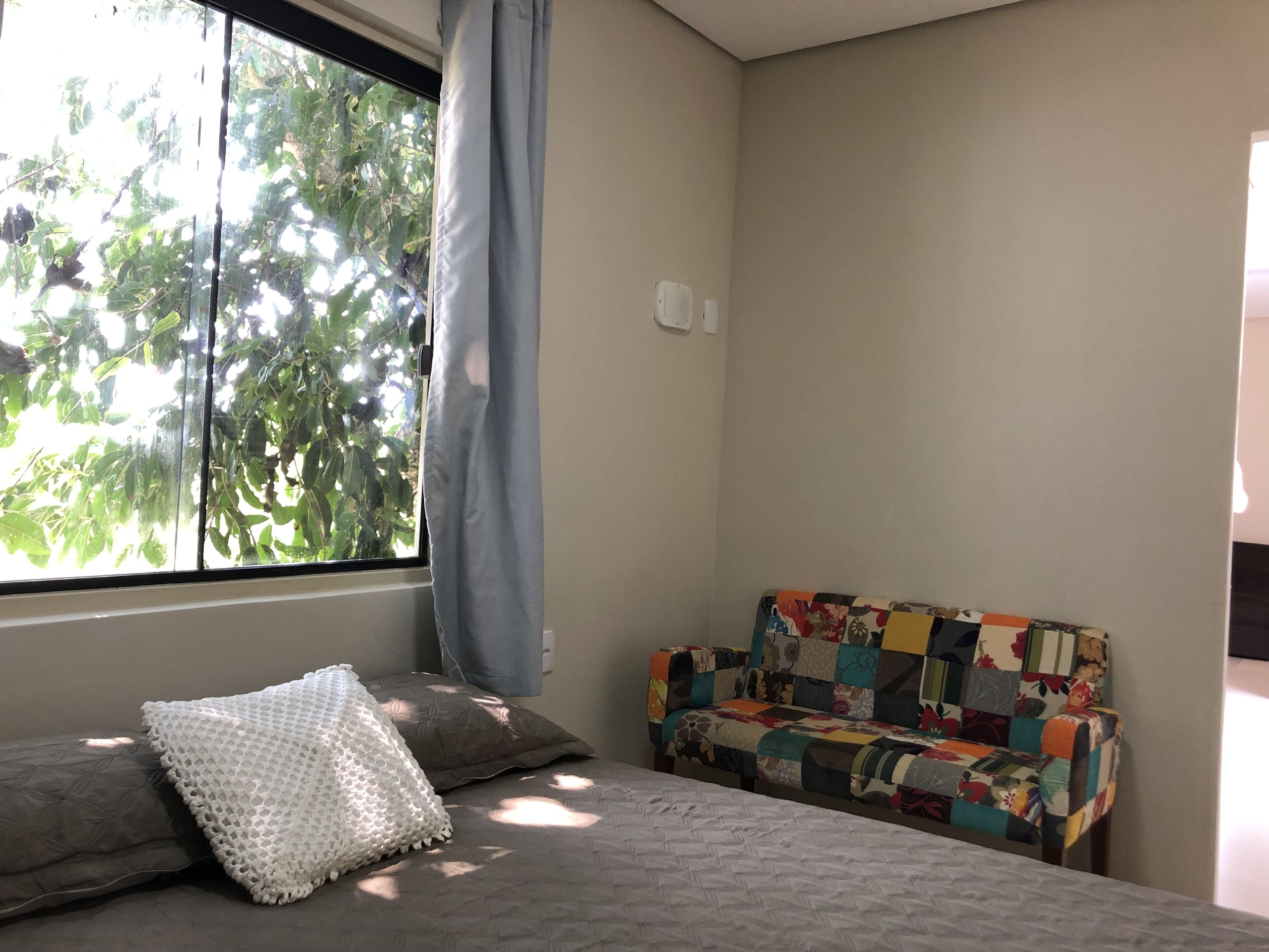 A bedroom with a bed, a colorful sofa, a window with curtains, and a view of trees outside.