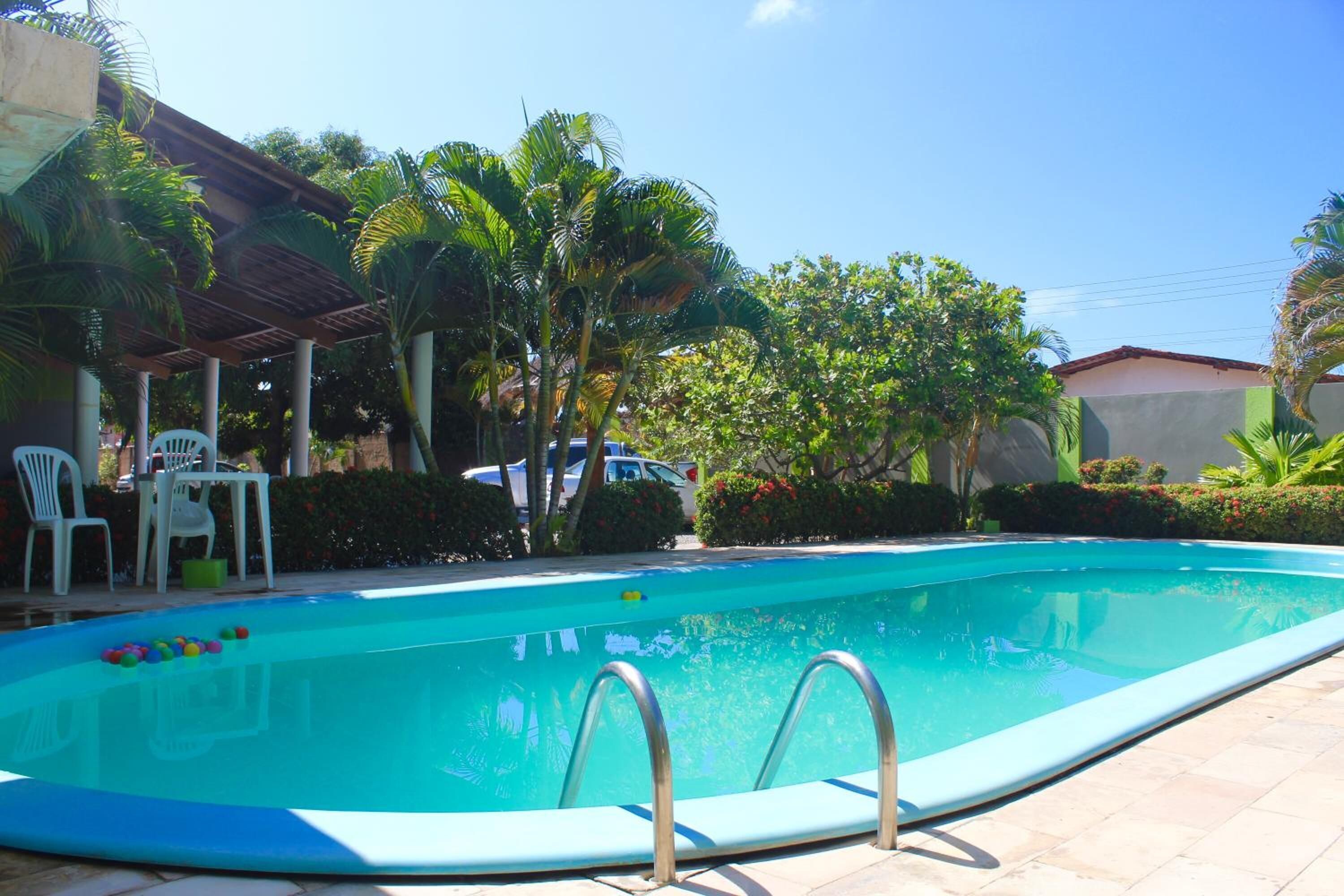 Piscina con escaleras de metal, rodeada de palmeras y una zona de estar cubierta con sillas blancas.