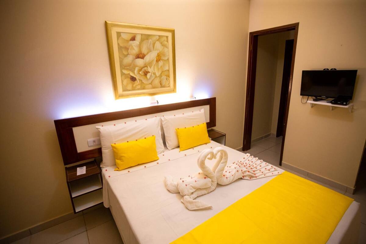 A hotel room with a bed, a painting above the bed, a television on a stand, and a door leading to another room.