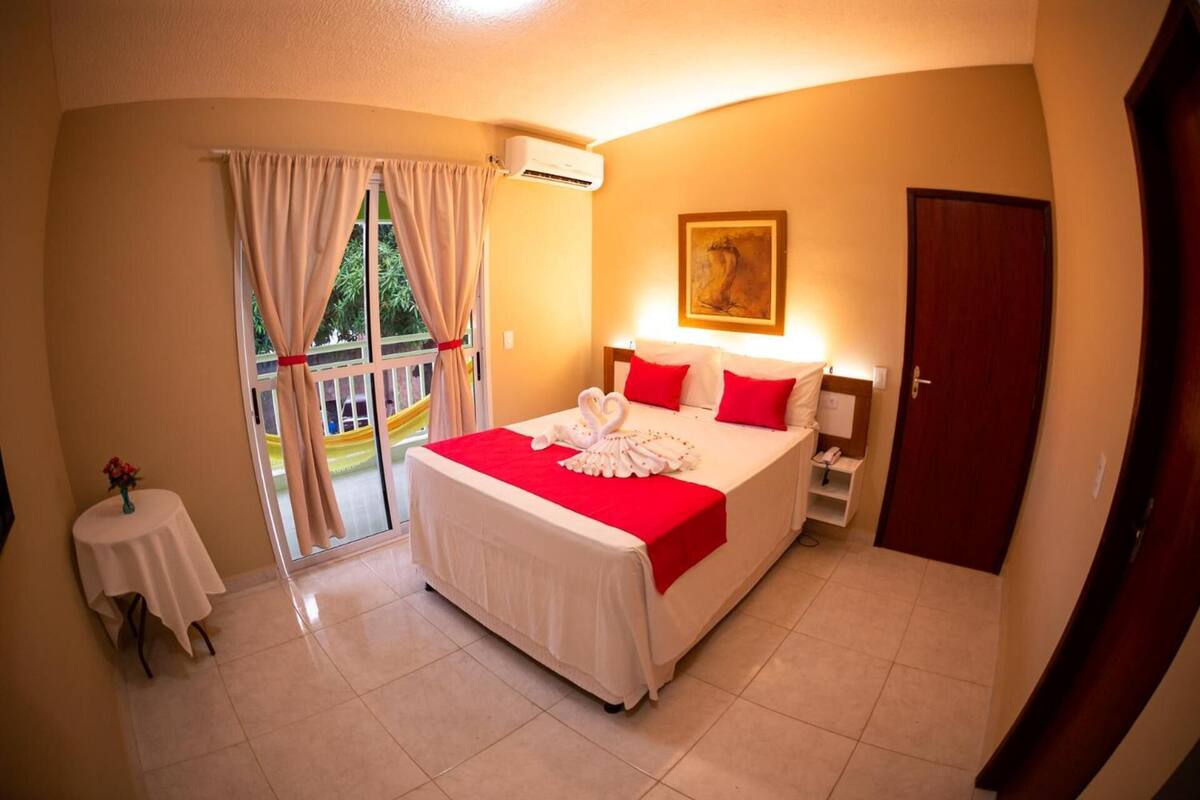 A hotel room with a bed, a small table with a vase, a window with curtains, and a door.
