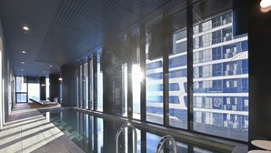 Una piscina interior moderna con paredes de vidrio transparente, piso de baldosas y barandas de metal.