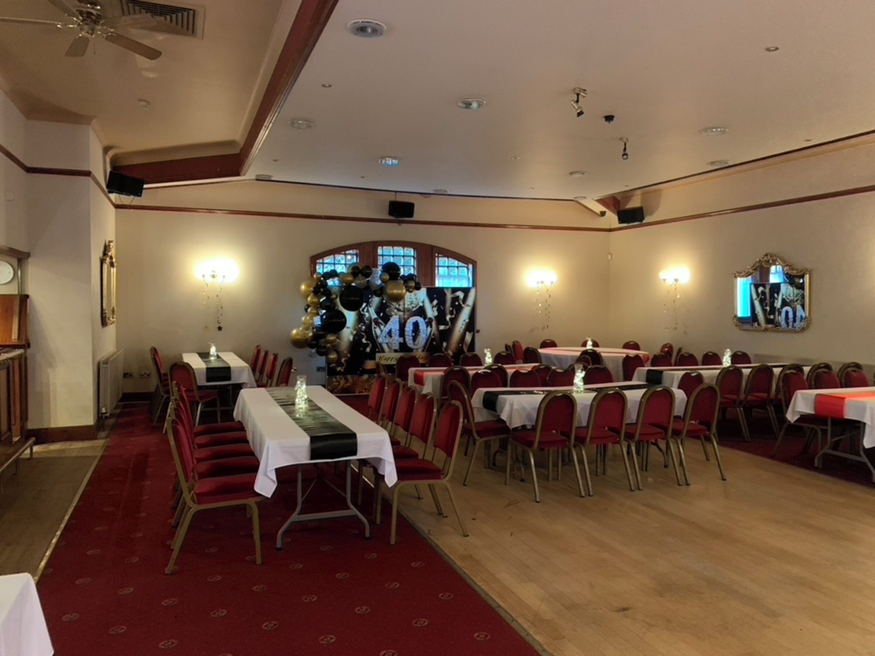Un salón de banquetes con varias mesas redondas, sillas rojas y un escenario con globos.