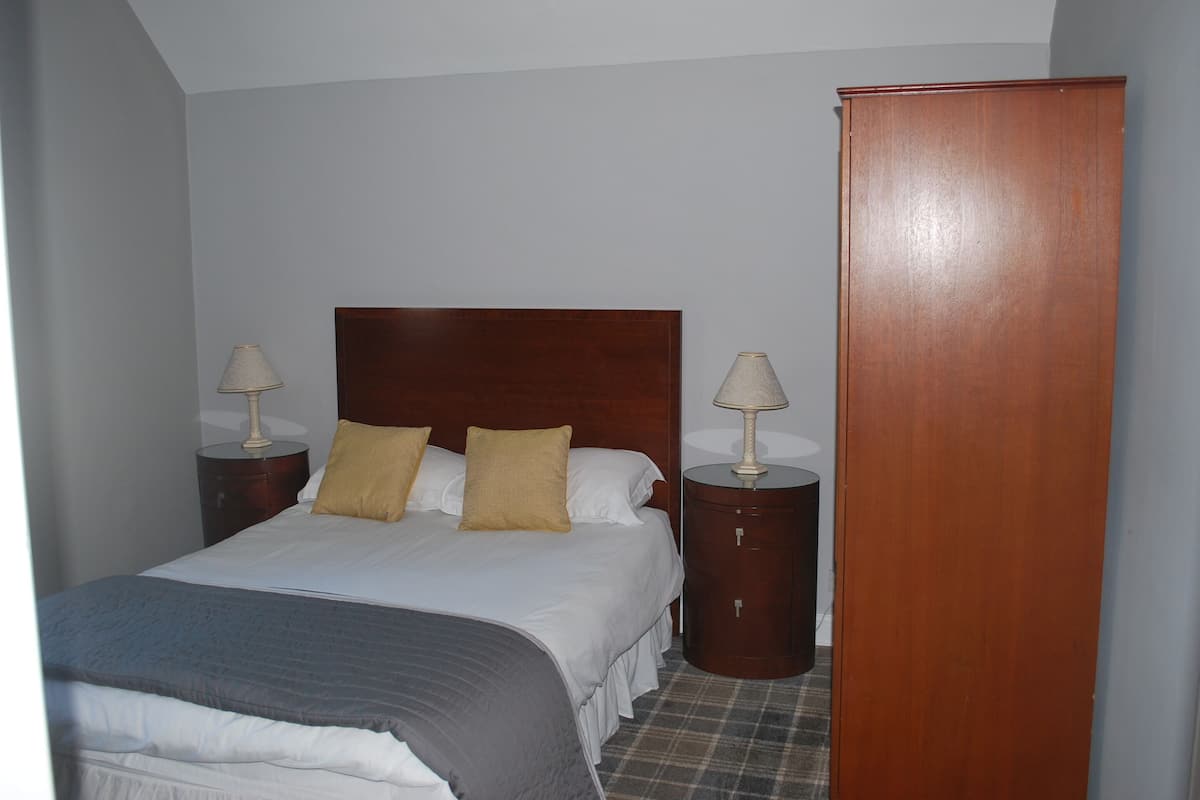 A neatly made bed with white and gray bedding, two bedside lamps, and a wooden wardrobe in a room with light-colored walls and a plaid floor.