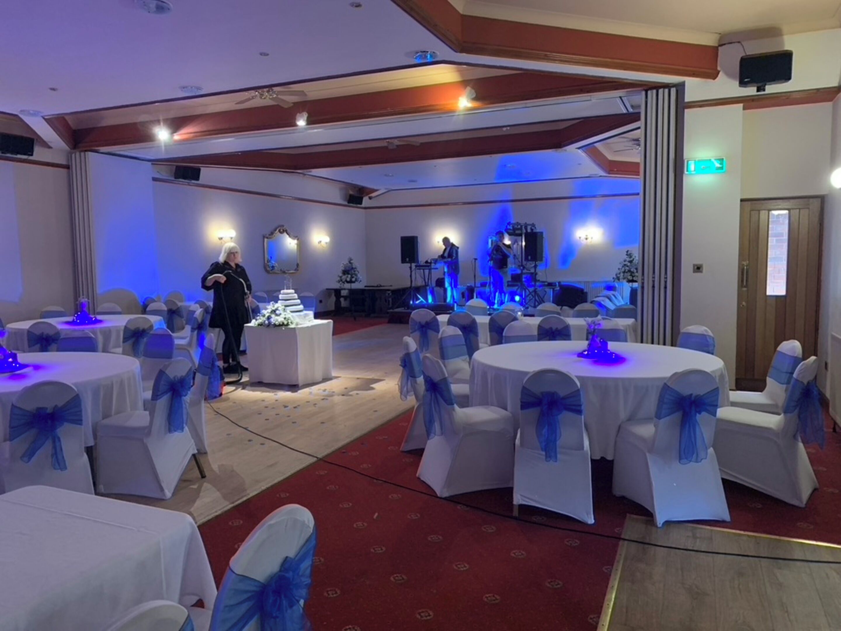 A banquet hall with round tables covered in white tablecloths and blue sashes, a stage with a band, and a red carpet.