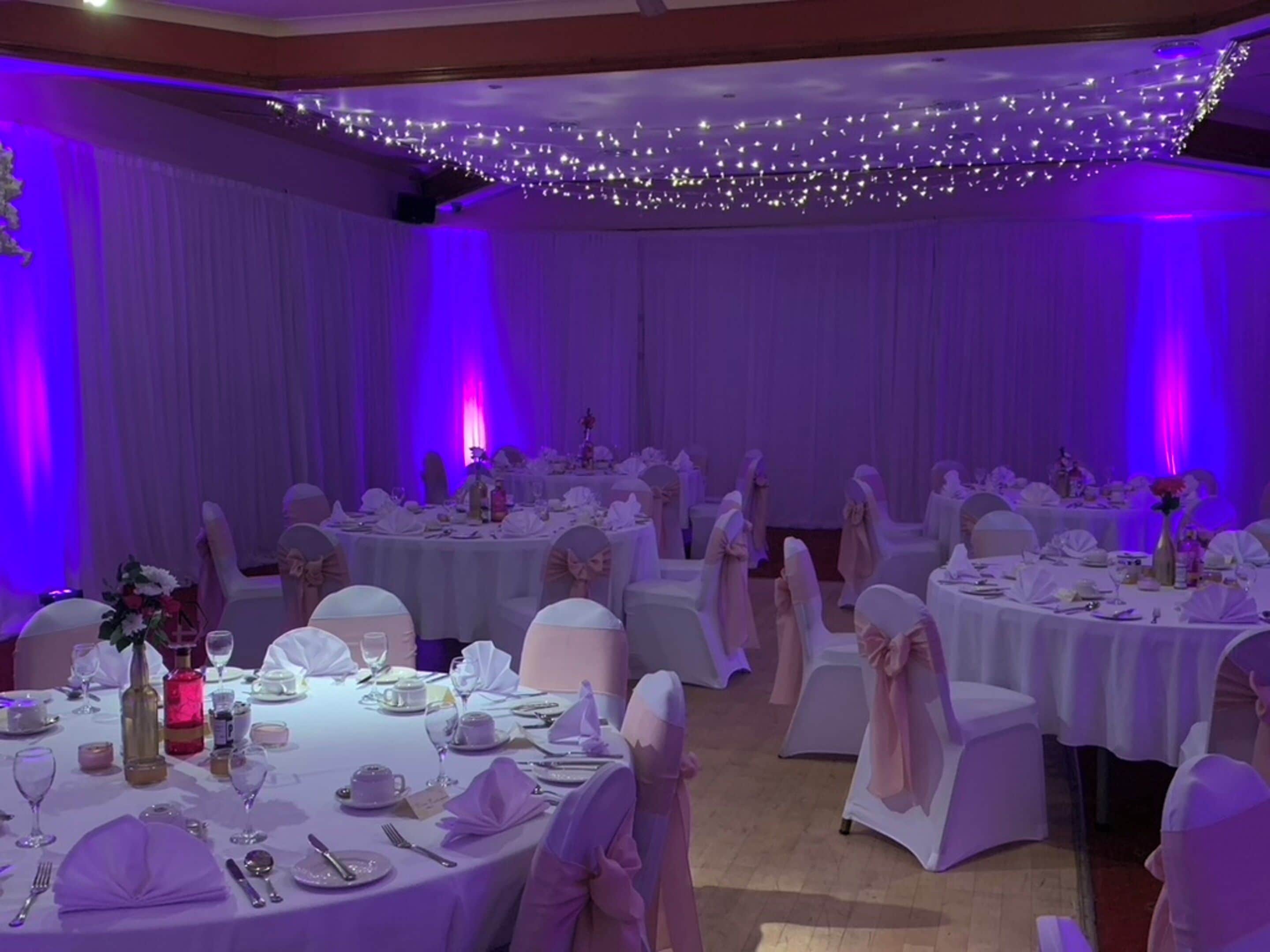 Um salão de banquetes com mesas redondas dispostas para um evento formal, com toalhas brancas, cadeiras com laços e teto decorado com luzes de Natal.