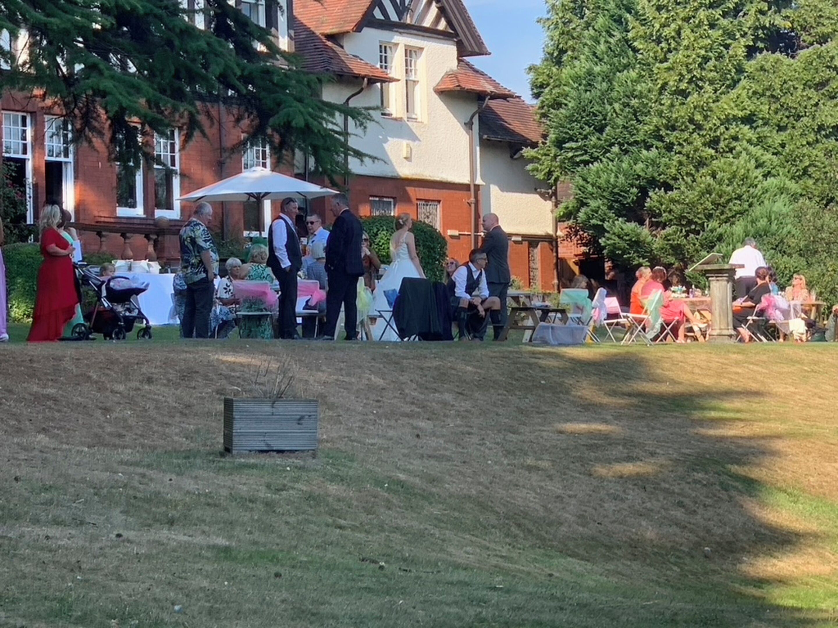 An outdoor gathering with people seated at tables, a grassy area, and a building with a red roof in the background.