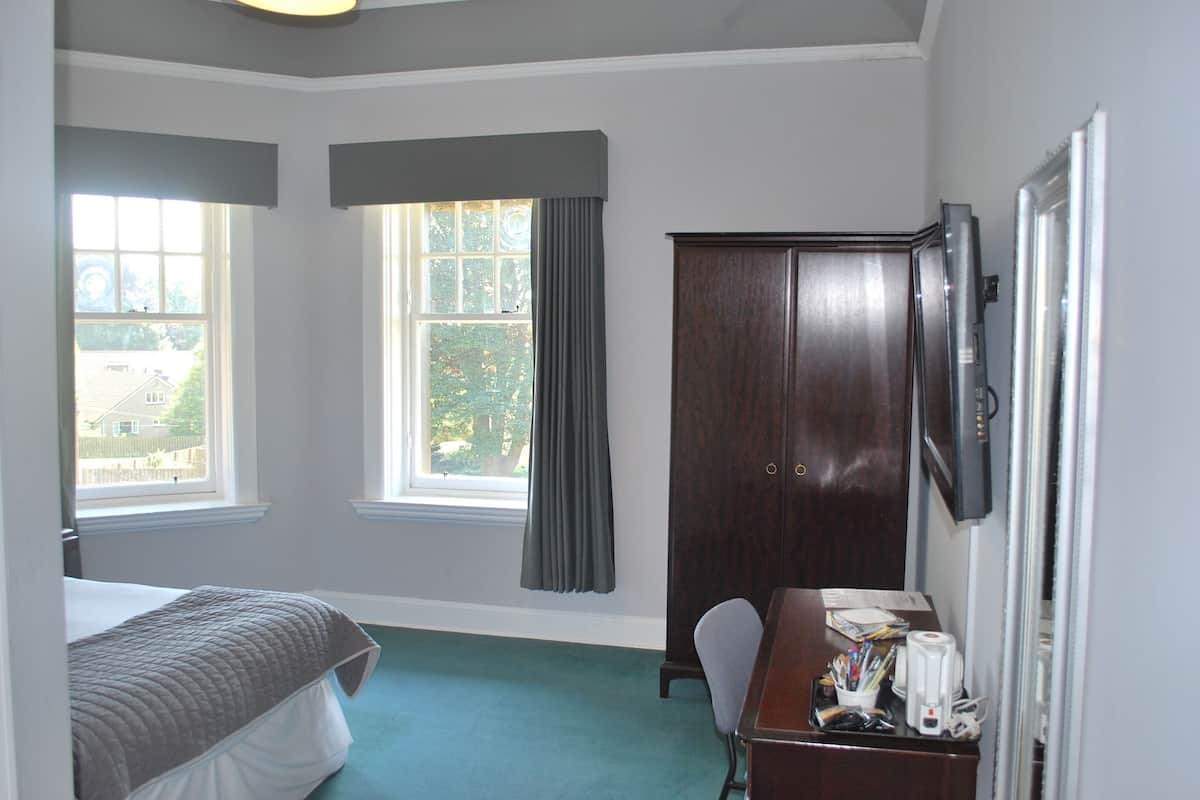 A bedroom with a bed, a desk, a chair, a wardrobe, and two windows with curtains.