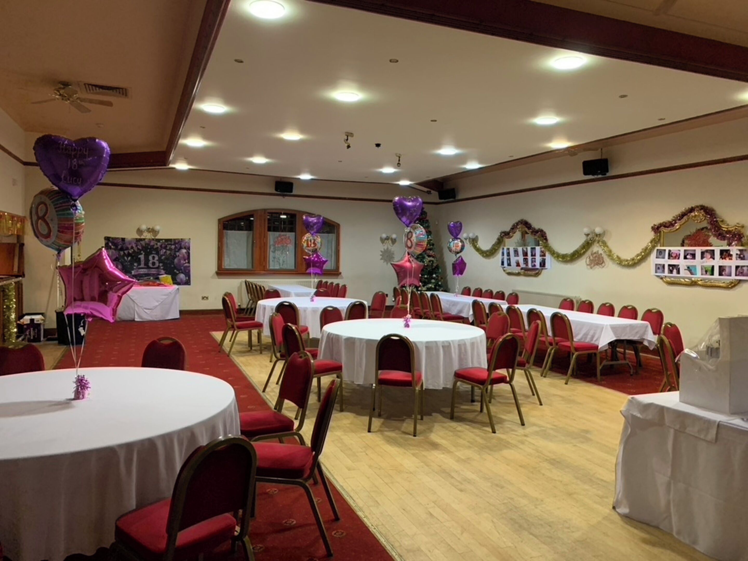 A banquet hall with round tables and red chairs, decorated with balloons and a Christmas tree.