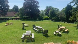 Un espacio al aire libre con césped, mesas y sillas de picnic, rodeado de árboles y un edificio al fondo.