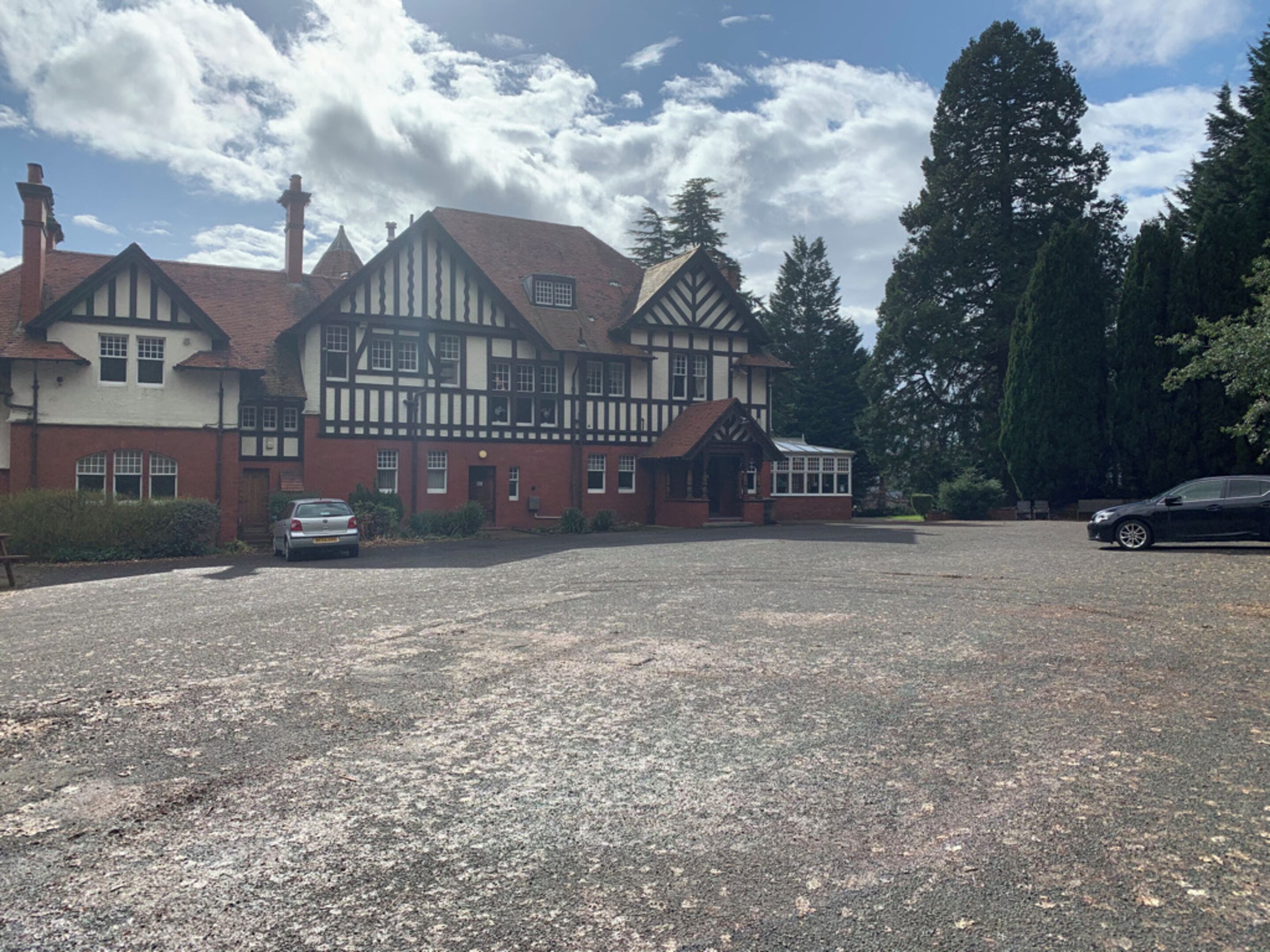 Un edificio grande de estilo Tudor de dos pisos, con camino de grava y coches aparcados.