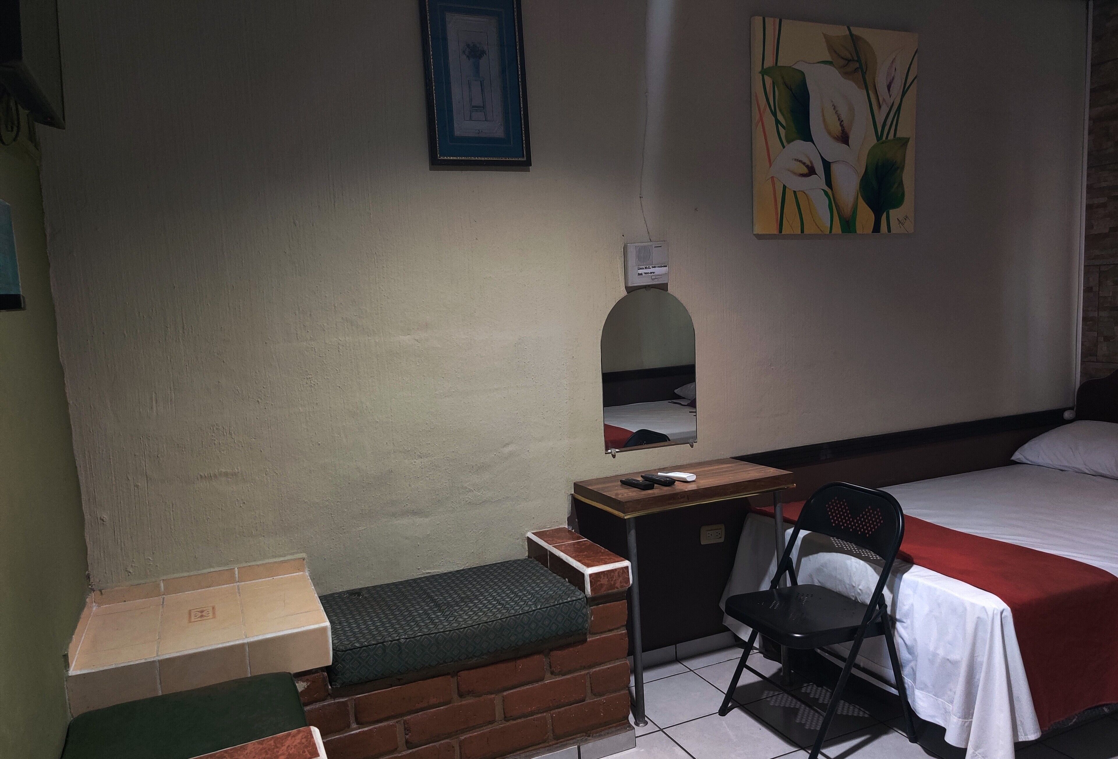 A hotel room with a bed, a desk, a chair, a small table, a bench, a picture on the wall, and a painting.