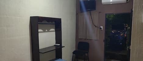 A hotel room with a bed, a desk with a chair, an air conditioning unit, a television, and a window with curtains.