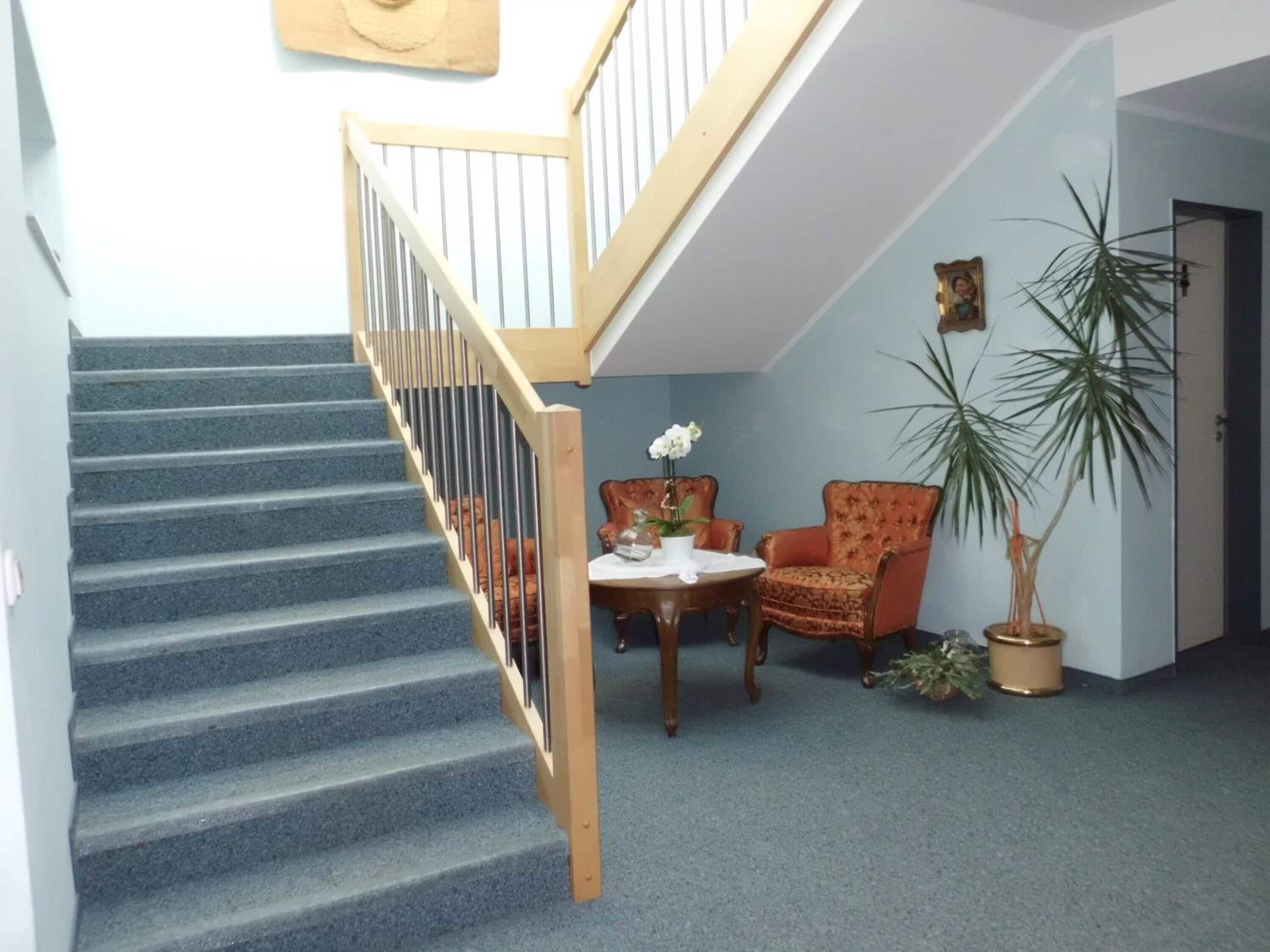 Una escalera con barandillas de madera que conduce a un piso superior. En la zona inferior, hay una mesa redonda con una planta en maceta y una silla.