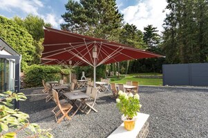 Un patio exterior con una sombrilla roja, mesas y sillas de madera y suelo de grava.