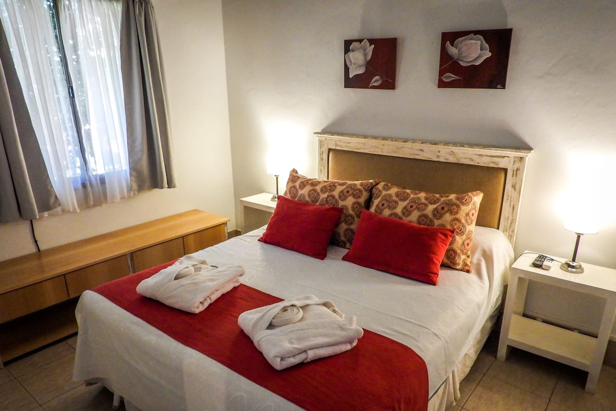 Un lit bien fait, avec une literie rouge et blanche, deux oreillers et deux serviettes pliées posées sur le lit. On y trouve deux tables de chevet avec des lampes, un banc en bois et deux tableaux accrochés au mur.
