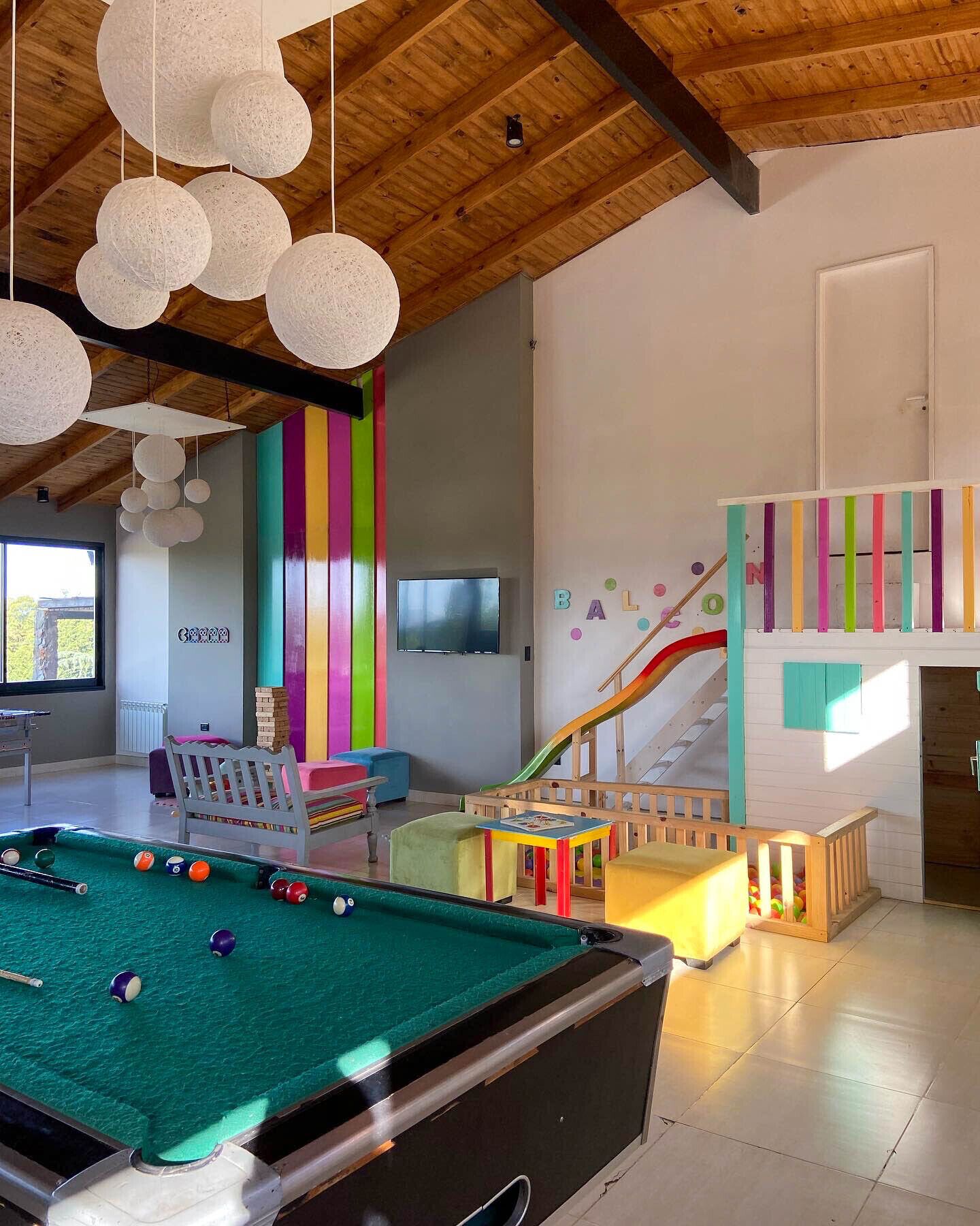 A modern indoor space with a pool table, colorful vertical stripes on the wall, a children's play area with a slide, and a staircase.