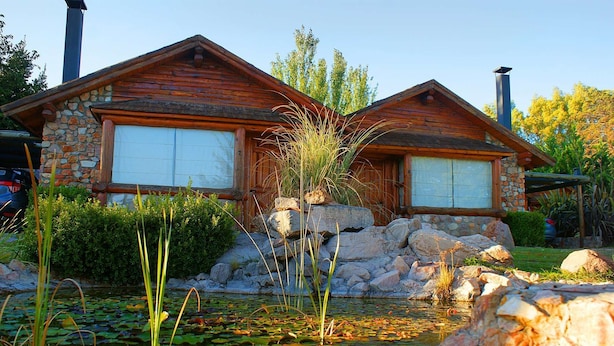 A rustic cabin with a stone foundation, wooden walls, and a pond with lily pads. - Balcon del Golf Resort, Spa & Restaurant. (Sierra de la Ventana)