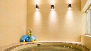 Ein Wellnessraum mit einer großen, runden Whirlwanne, beige gefliesten Wänden und drei Wandleuchten.