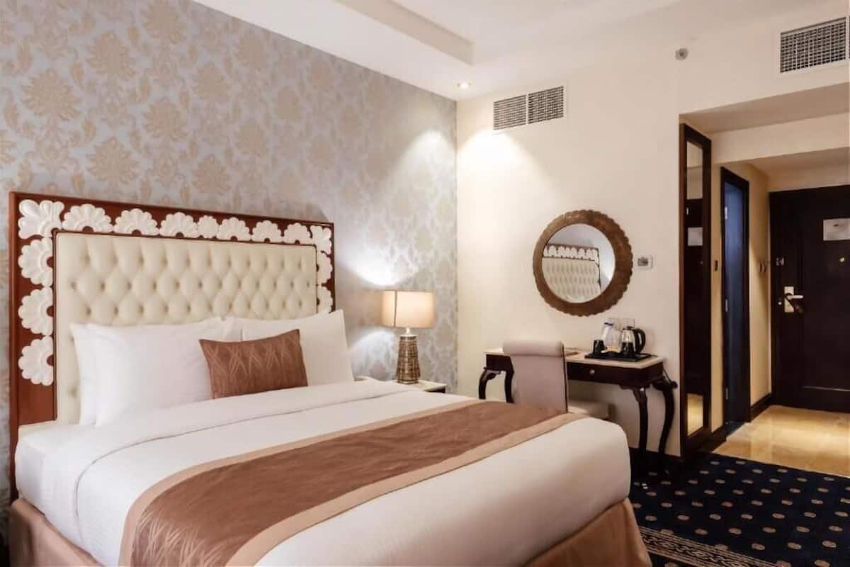 Una habitación de hotel con una cama grande, una mesita de noche con lámpara, un escritorio pequeño y un espejo en la pared.