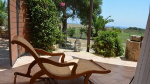 A wooden rocking chair with a woven seat and backrest, positioned on a tiled patio with a view of a lush garden and distant hills. - Alekos Beach Houses - Natura Rossa (Kefalonia)