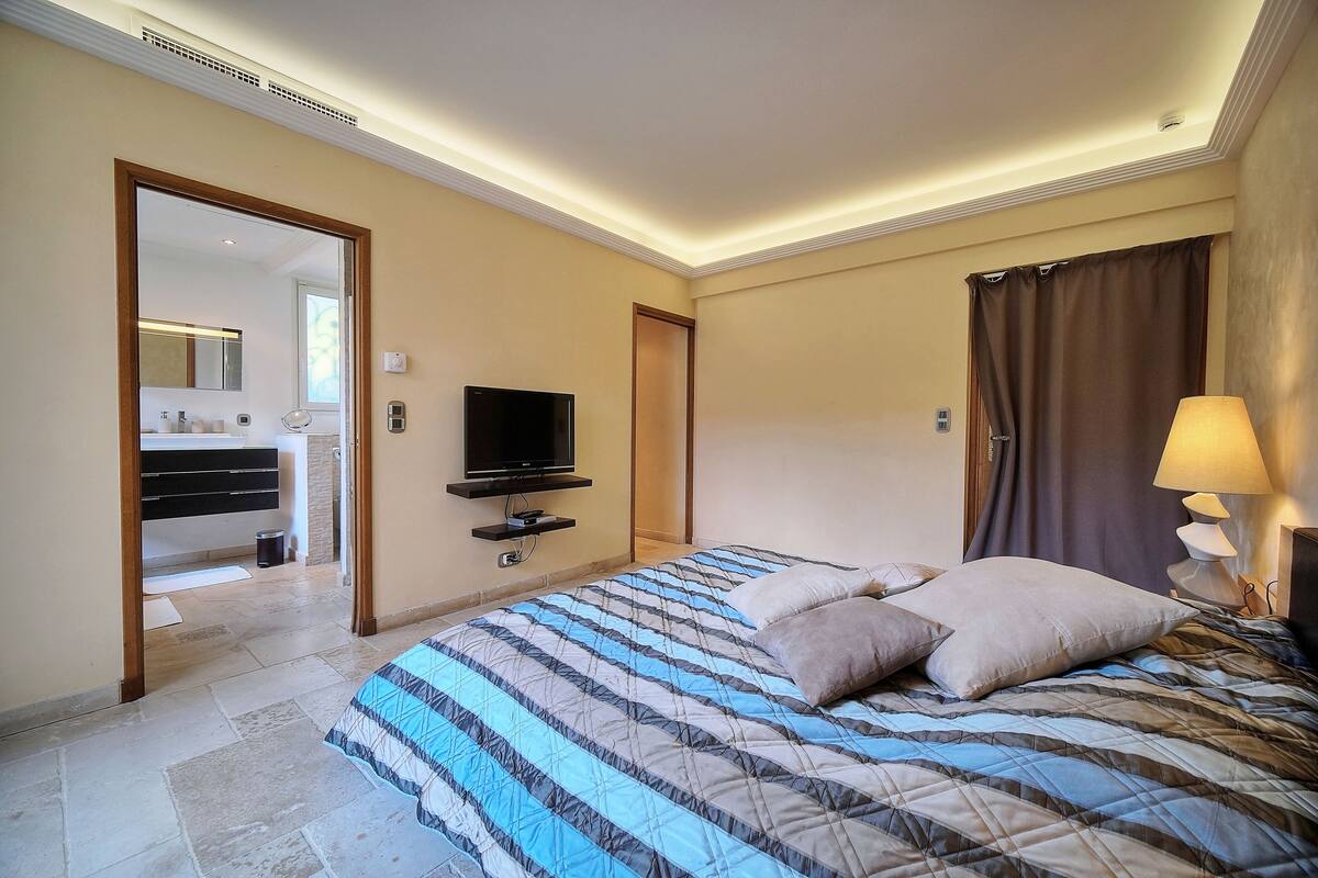 A hotel room with a bed, a television, a bathroom visible through an open door, and a nightstand with a lamp.