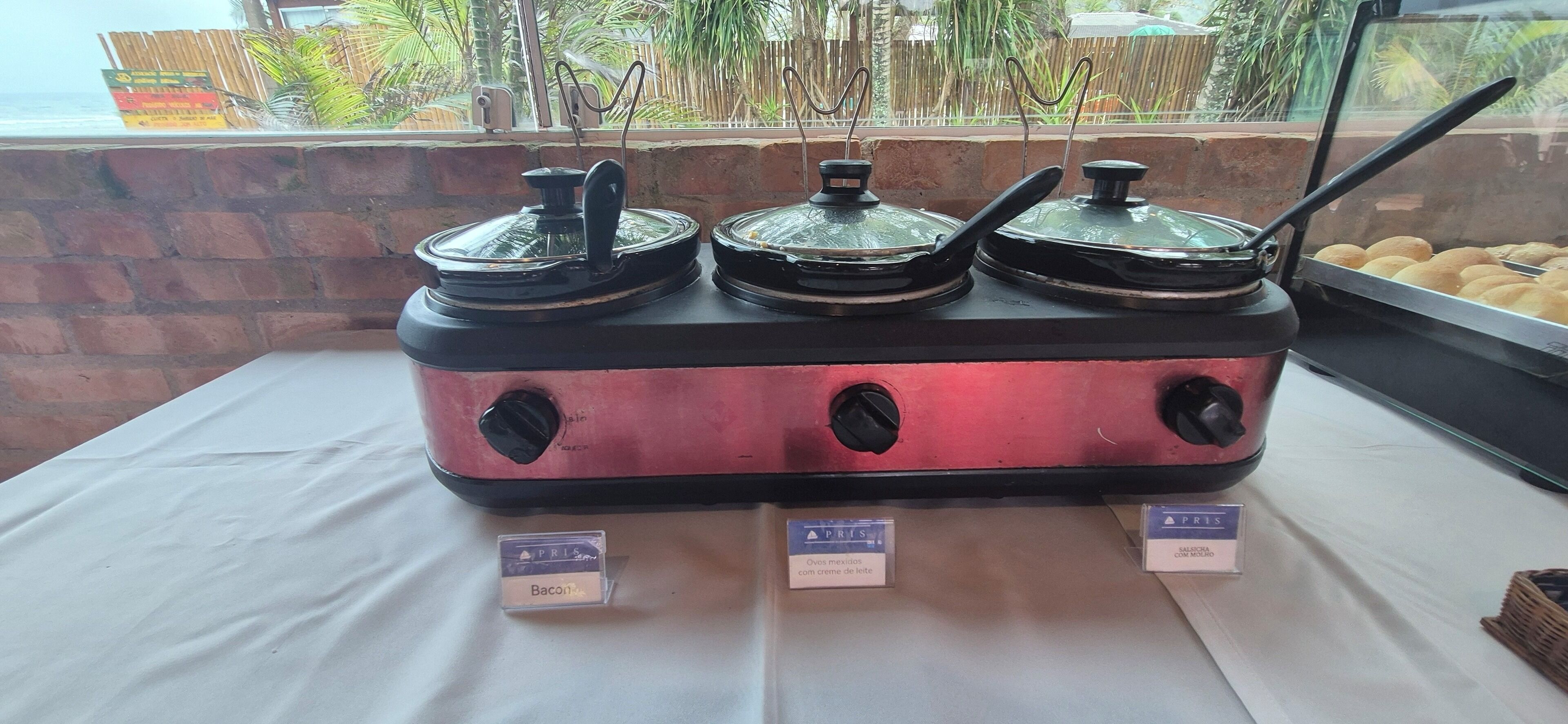 Montaje de bufé con tres dispensadores de comida cubiertos, etiquetados como 'Bacon', 'Ovos mexitos con crema de leche' y 'Saborosa con leche'.