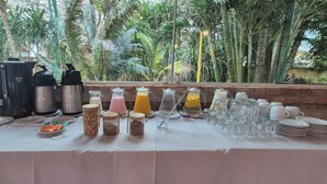 A breakfast buffet table with various food items and beverages, set against a backdrop of lush greenery and a brick wall. - PRIS Hotel (São Sebastião)