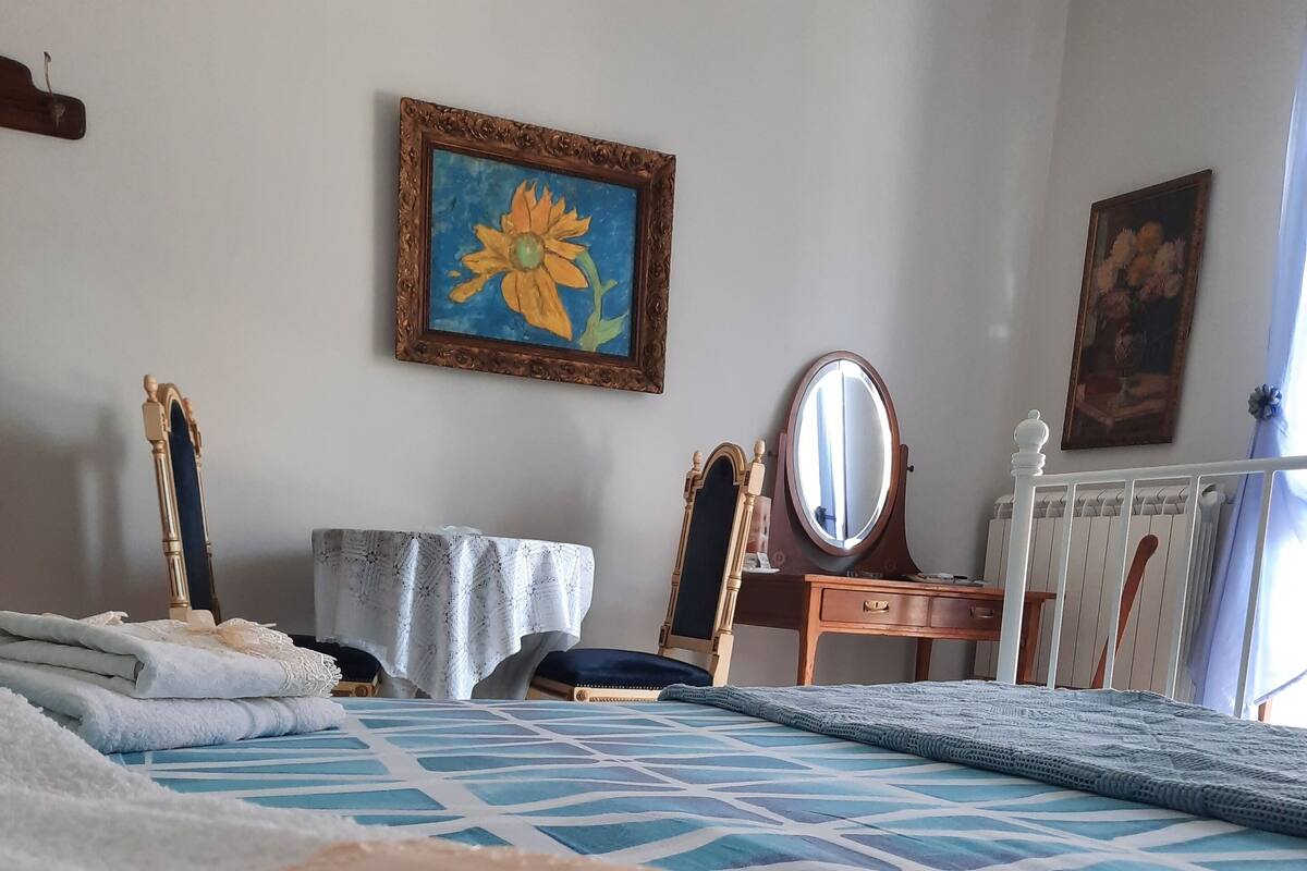 A neatly made bed with a blue and white patterned bedspread, a wooden chair with a cushion, a small table with a lace tablecloth, and a wall with two framed paintings.