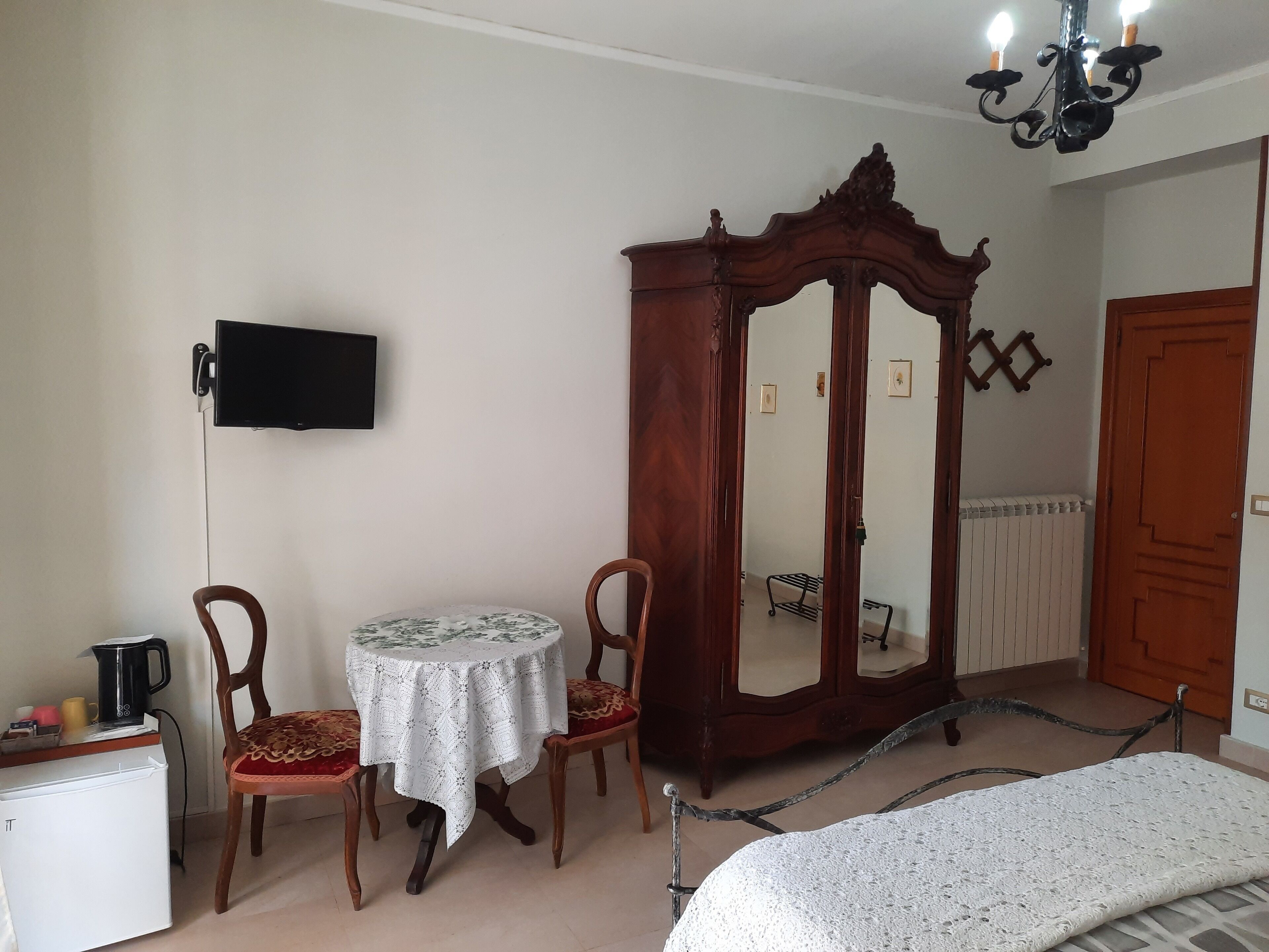 A bedroom with a bed, a wooden wardrobe, a round table with chairs, a television mounted on the wall, and a radiator.