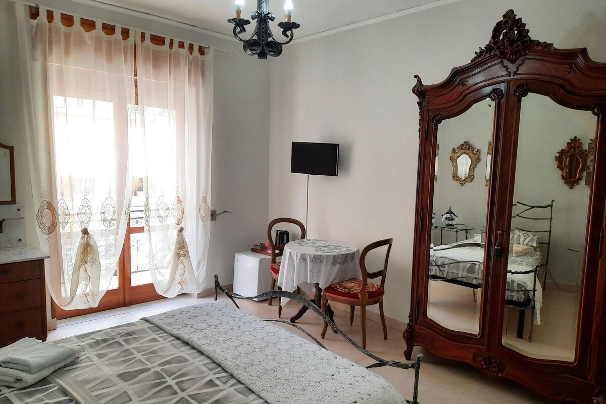 A bedroom with a bed, a wooden wardrobe, a desk with a chair, a television, and a large mirror.