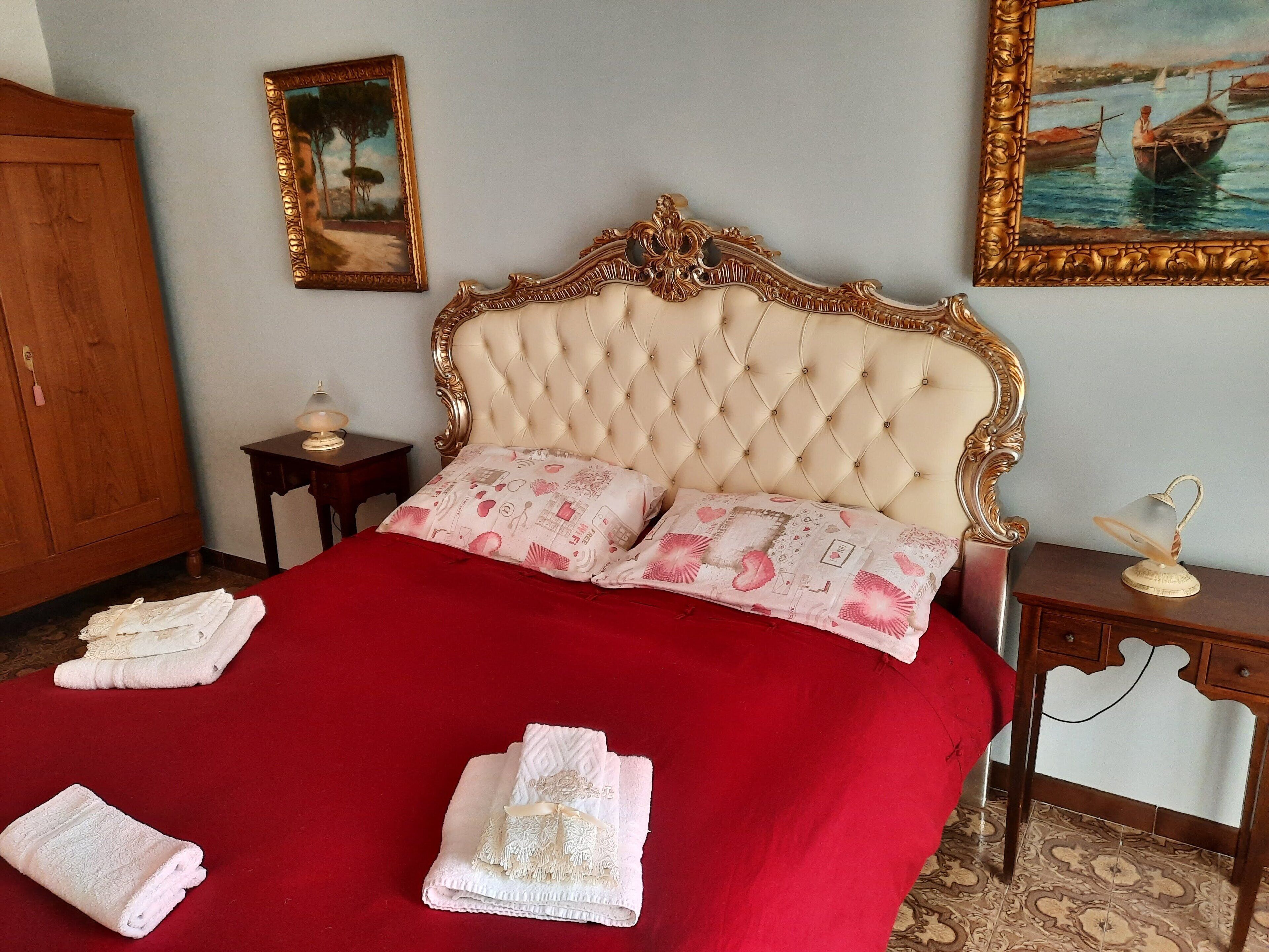 Un dormitorio con cabecera capitonada, dos almohadas con estampado floral, una colcha roja y dos toallas dobladas sobre la cama. Hay dos mesitas de noche, una con una lámpara y la otra con un adorno.