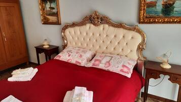 Un dormitorio con cabecera capitonada, dos almohadas con estampado floral, una colcha roja y dos toallas dobladas sobre la cama. Hay dos mesitas de noche, una con una lámpara y la otra con un adorno.