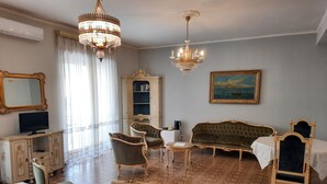 A room with a large ornate chandelier, a green upholstered sofa, a wooden chair, a small table, a cabinet with a mirror, and a painting on the wall. - Villa Ada B&B (Agropoli)