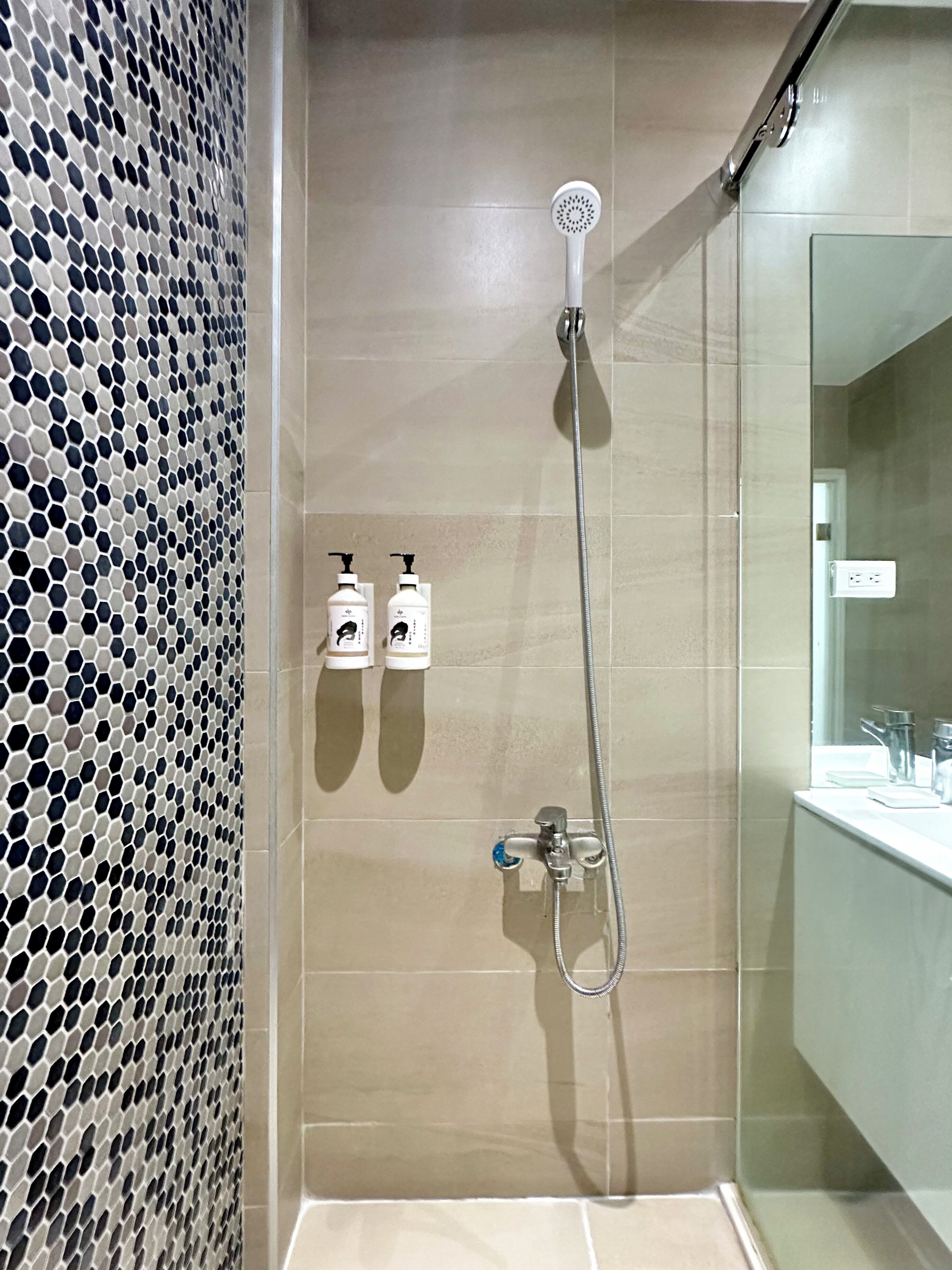Una doccia con soffione a mano, rubinetto montato a parete e due flaconi di shampoo e balsamo.