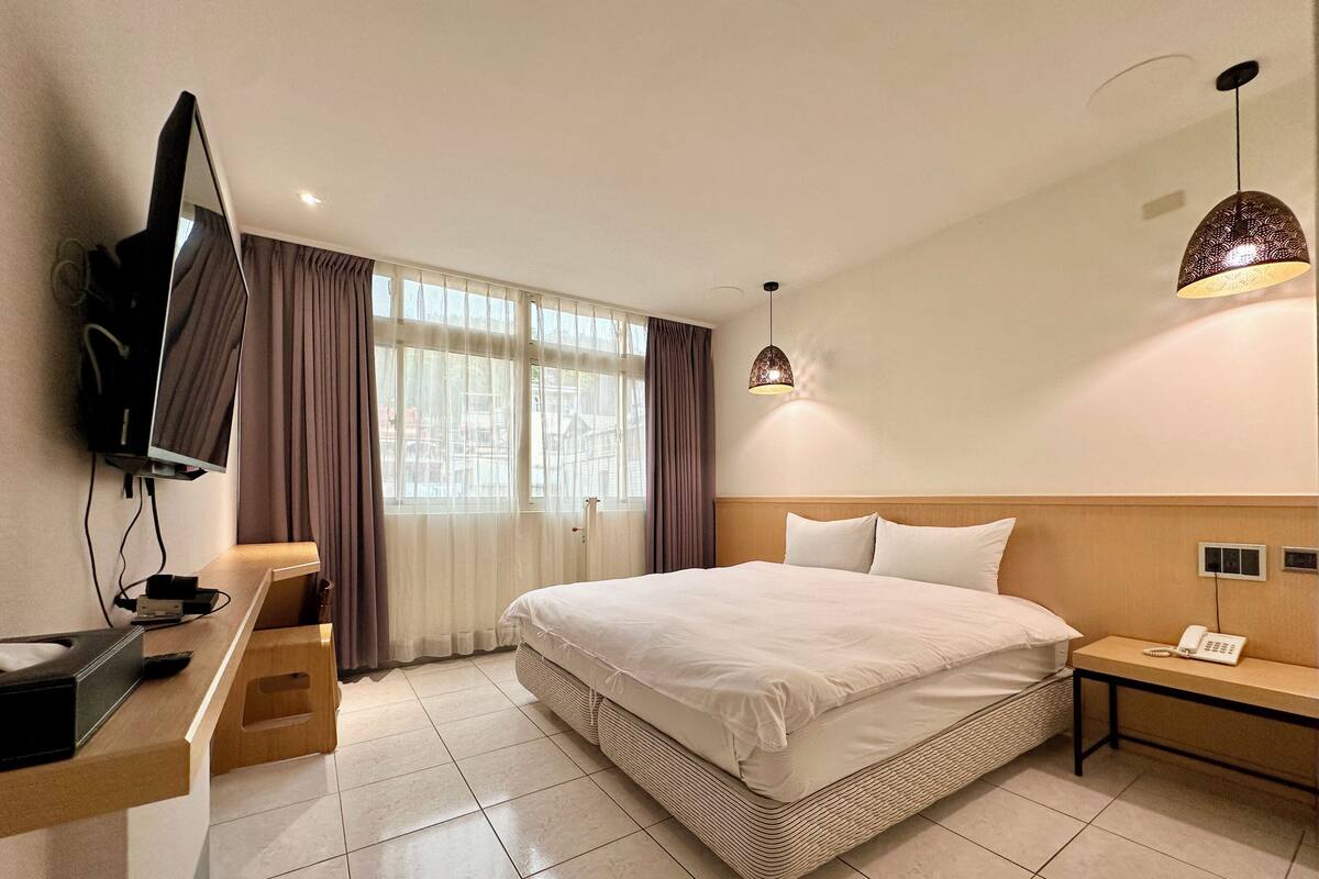 A hotel room with a bed, a desk with a TV, a bedside table with a lamp, and a window with curtains.