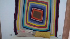 Quarto com cama, almofadas, um cobertor e um pendente de parede colorido.