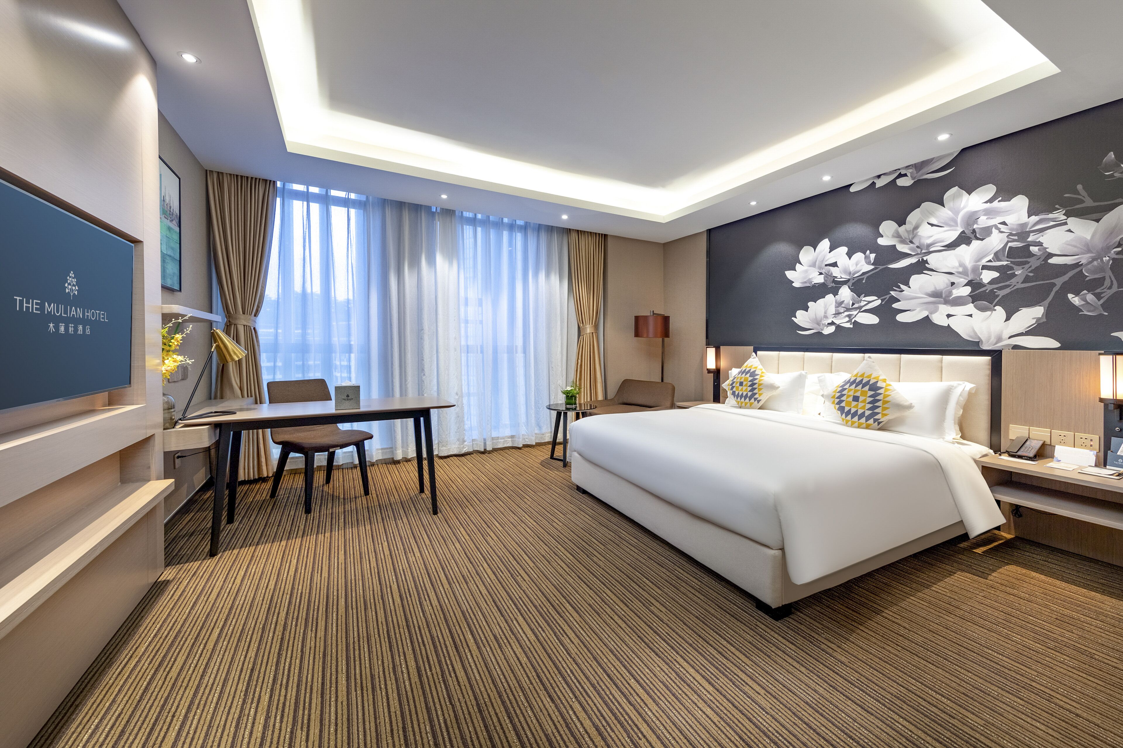 A hotel room with a bed, a desk, a chair, a TV, and a wall with a floral mural.
