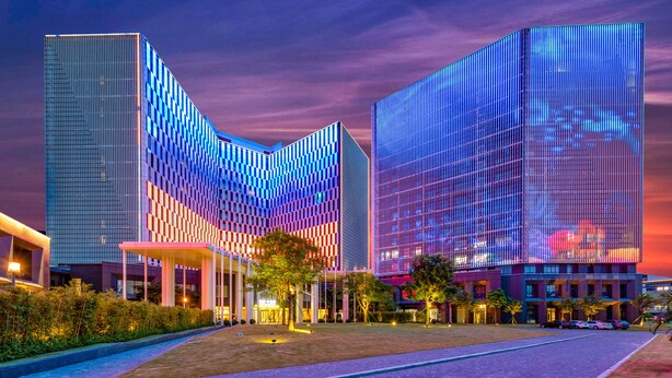 A modern hotel with a distinctive curved facade, a spacious outdoor area with a paved walkway, and a well-lit entrance.