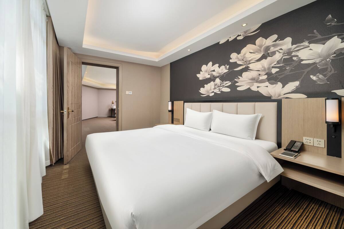 A hotel room with a large bed, a nightstand with a phone and a lamp, a floral wall mural, and a door leading to another room.