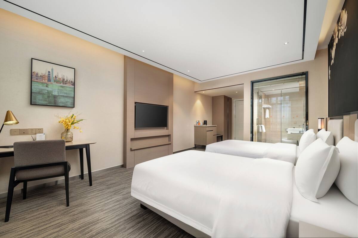 A hotel room with two beds, a desk, a TV, and a bathroom visible through a glass door.