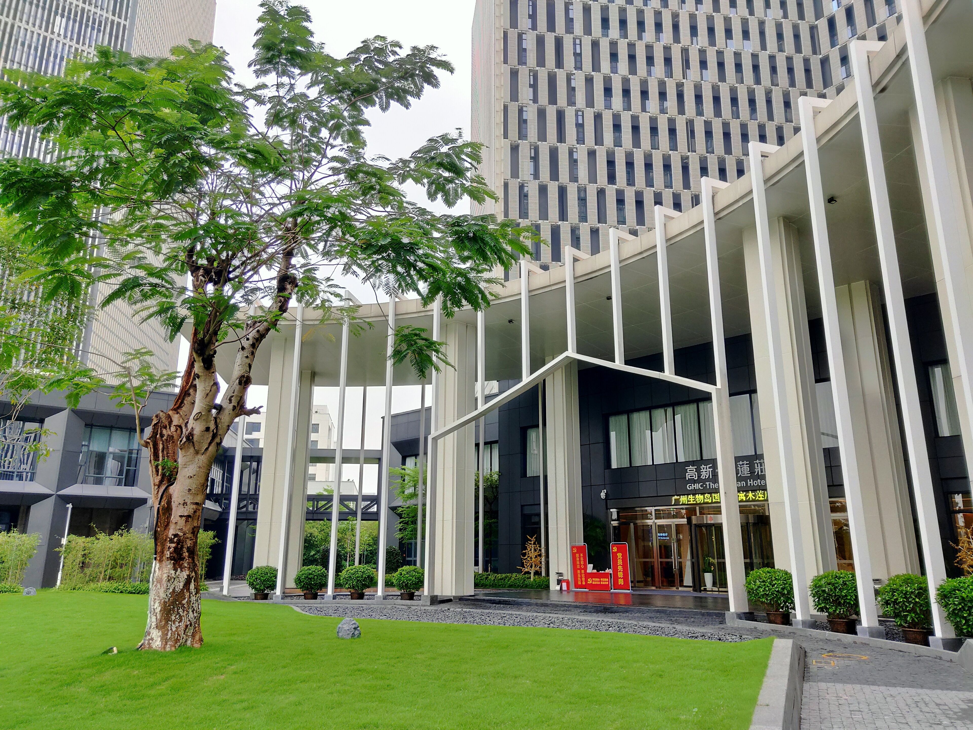 Un edificio moderno con una entrada amplia, rodeado de vegetación y un árbol.