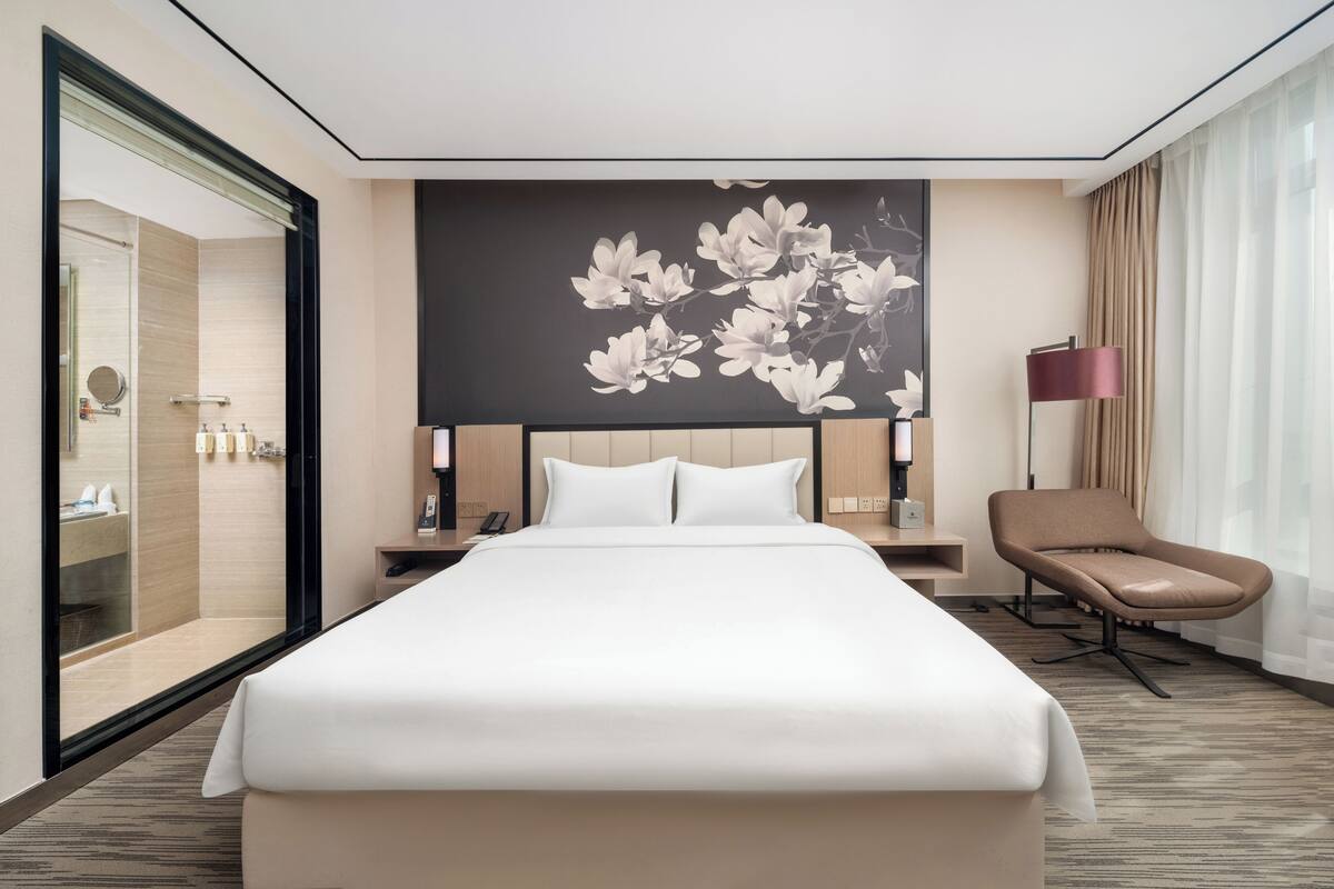 A hotel room with a bed, a chair, a nightstand, a lamp, and a wall with a floral design.