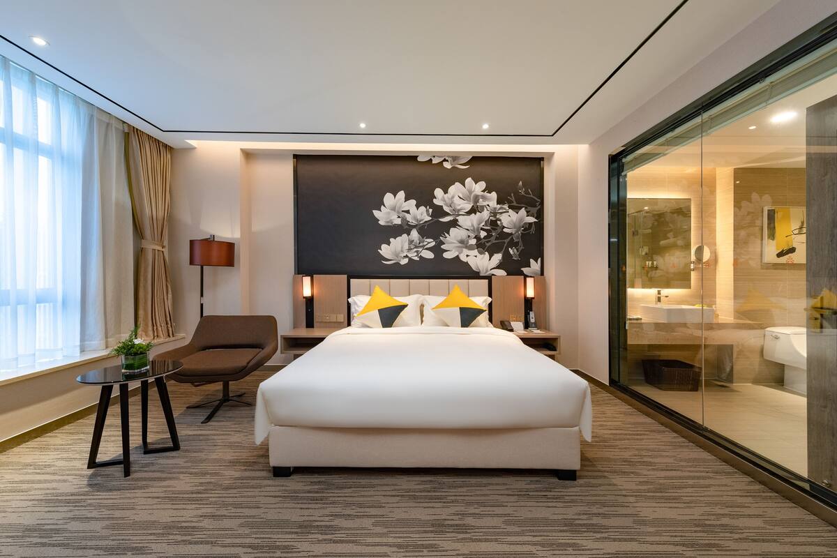 A modern hotel room with a large bed, a seating area with a chair, a small table with a plant, a large wall-mounted artwork, and a glass-enclosed bathroom.