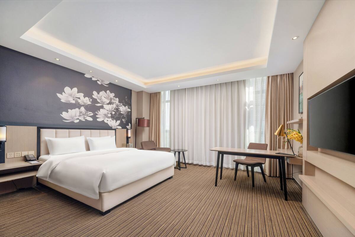 A hotel room with a large bed, a desk, a chair, a TV, and a wall with a floral design.