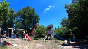 Un parque infantil con tobogán y columpios, rodeado de árboles y una casa al fondo.