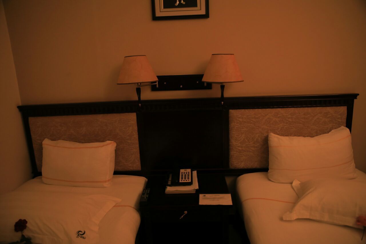 Une chambre d’hôtel avec deux lits, chacun étant équipé d’un oreiller et d’une couverture. Il y a une table de chevet entre les lits, sur laquelle se trouvent un téléphone et un petit objet.
